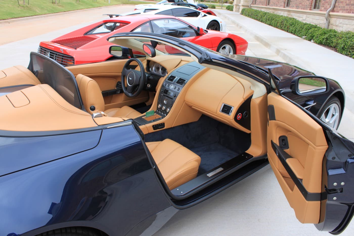 The freshly detailed tan leather cockpit of an Aston Martin V8 Vantage Roadster, door open, with a Ferrari Testarossa parked behind